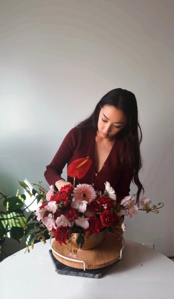 A florist in a burgundy top arranging a red anthurium and carnation centerpiece