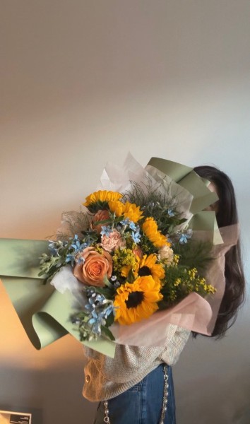 A person holding a sunflower and orange-rose bouquet wrapped in sage-green paper